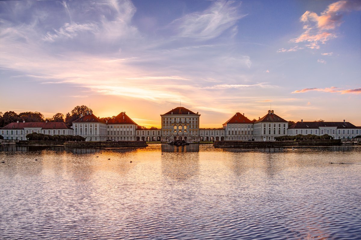 The photo shows Nymphenburg Castle during sunset, in the foreground there is a body of water