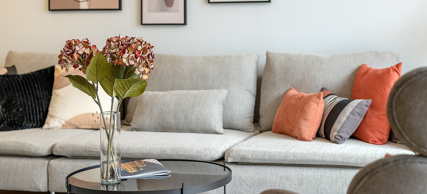 You can see the backrest of a sofa and a small glass table with flowers in a vase in front of it