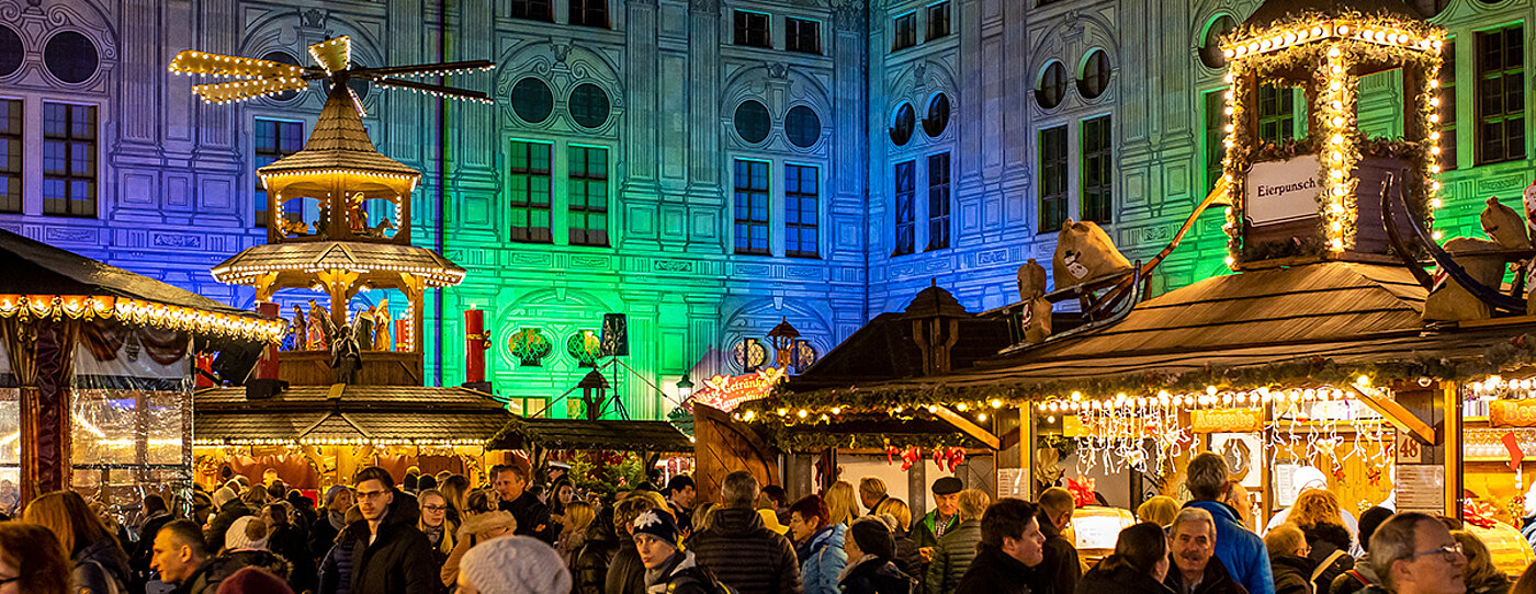 Zu sehen ist ein gut besuchter Christkindlmarkt, mit Ständen im Vordergrund und blau-grüner Beleuchtung im Hintergrund
