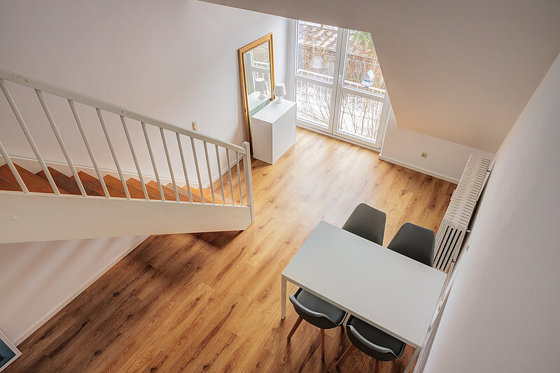 Vorher: Blick von oben in einen Wohnraum mit Holzboden, Treppe mit weißem Geländer und Esstisch mit zwei Stühlen 