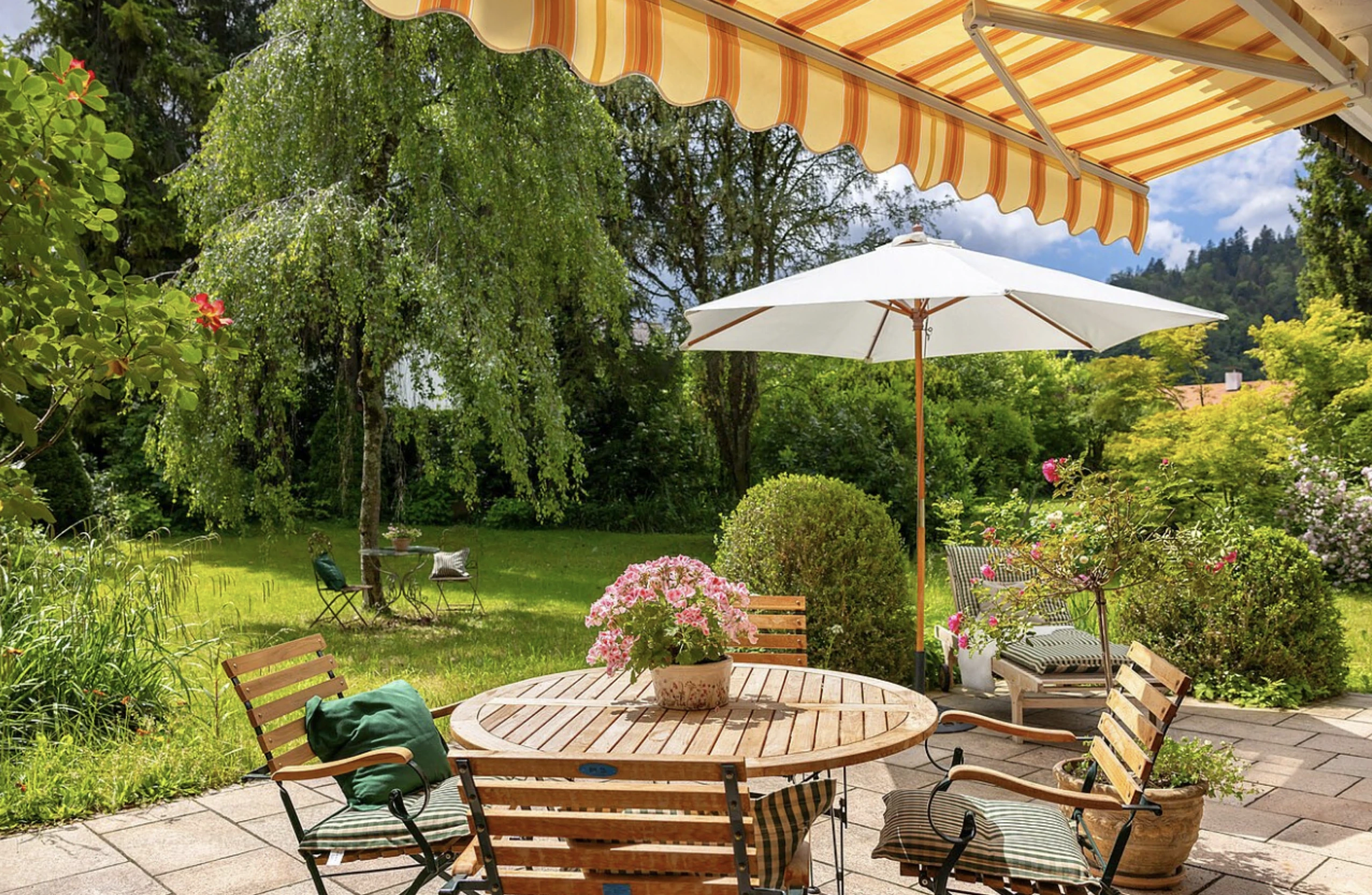 Gepflegte Gartenterrasse mit Holztisch, Sonnenschirm und gestreifter Markise vor blühendem Garten